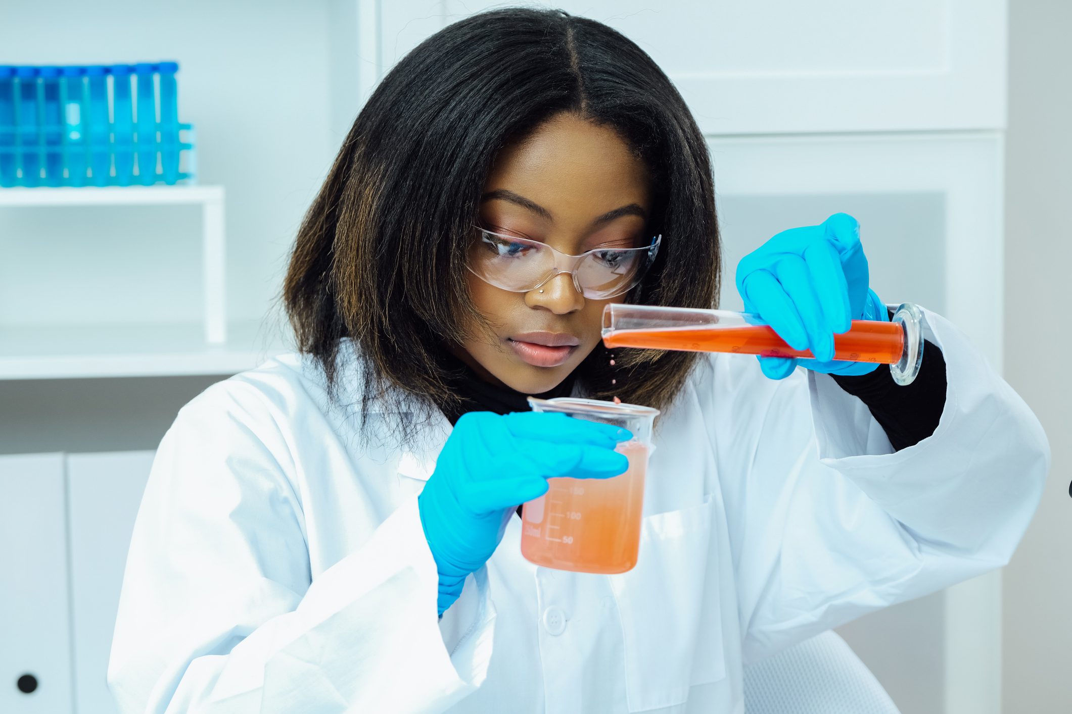 Scientist carefully pouring orange liquid in lab beaker, wearing gloves and goggles.